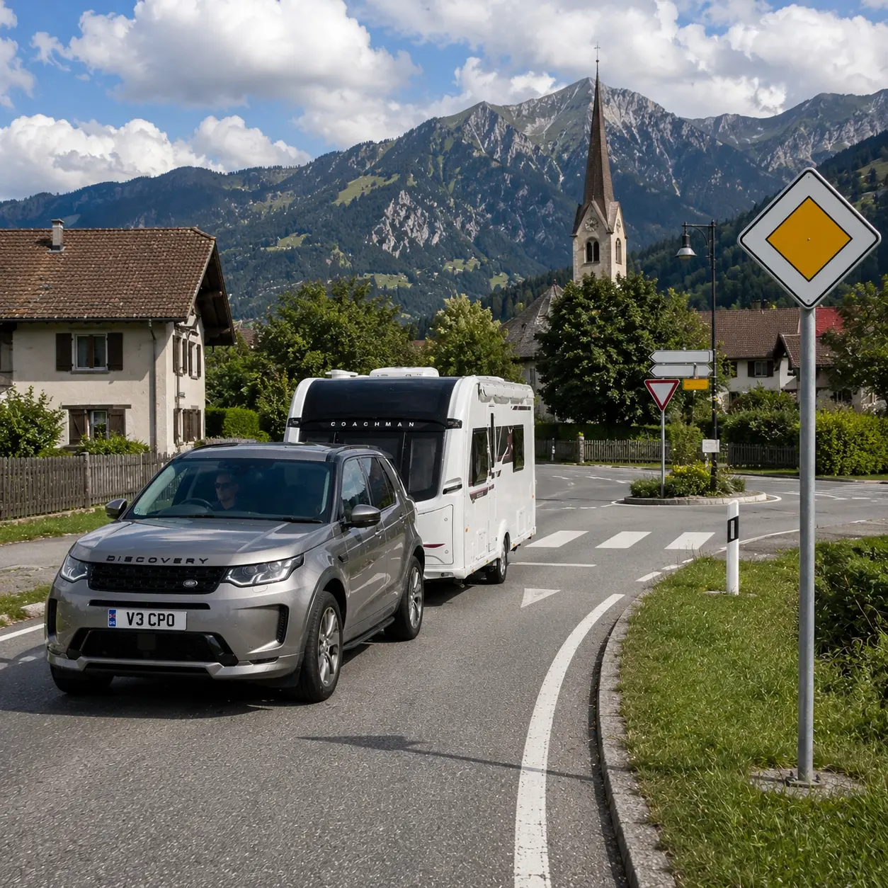 Silver Land Rover Discovery towing a Coachman caravan past a European priority road sign in an Alpine village